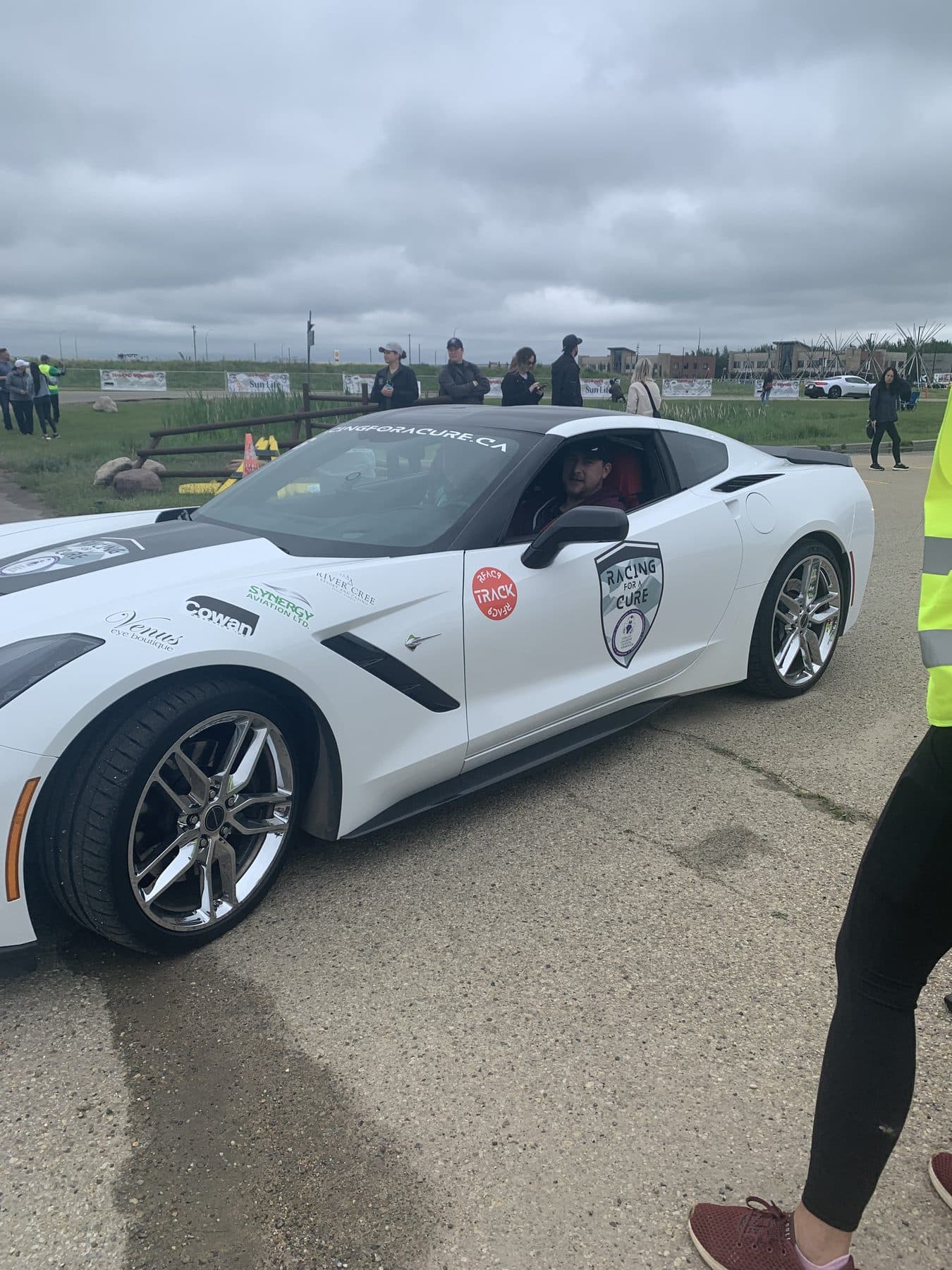 A white Corvette with Racing for a Cure decals beside a volunteer in a high-visibility vest.