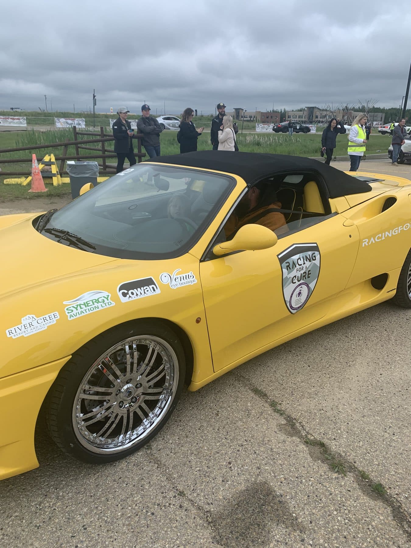 Side view of a yellow Ferrari covered in sponsor decals, including Racing for a Cure, at a charity event.